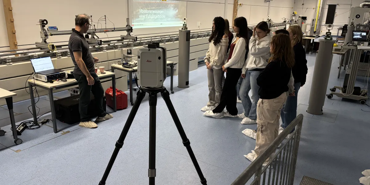 Herbstakademie 2025: Eine Gruppe Sch&uuml;lerInnen (ca. zwischen 16 und 18 Jahren) stehen in einem Laborraum. Ein Lehrender zeigt ihnen auf einem Testaufbau die statischen Eigenschaften von Br&uuml;cken.