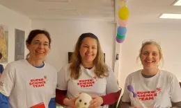 Drei Wissenschaftlerinnen in wei&szlig;en T-Shirts l&auml;cheln in die Kamera. Auf den Shirts sieht man den Schriftzug des Super Science Teams. In ihren H&auml;nden halten sie Flyer, Laborequipment und ein H&auml;febakterien-Stofftier.
