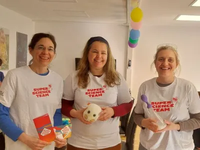 Drei Wissenschaftlerinnen in wei&szlig;en T-Shirts l&auml;cheln in die Kamera. Auf den Shirts sieht man den Schriftzug des Super Science Teams. In ihren H&auml;nden halten sie Flyer, Laborequipment und ein H&auml;febakterien-Stofftier.