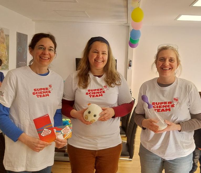 Drei Wissenschaftlerinnen in wei&szlig;en T-Shirts l&auml;cheln in die Kamera. Auf den Shirts sieht man den Schriftzug des Super Science Teams. In ihren H&auml;nden halten sie Flyer, Laborequipment und ein H&auml;febakterien-Stofftier.