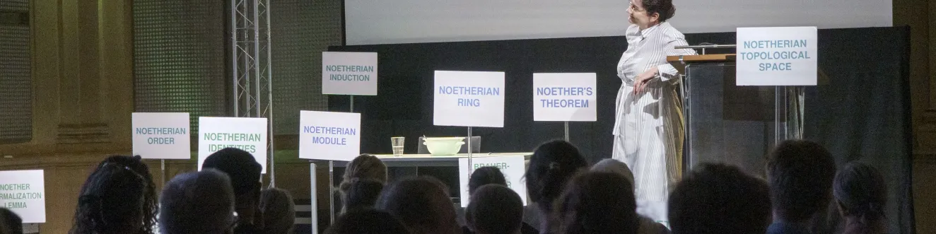 Foto vom Theaterst&uuml;ck "Diving into Math with Emmy Noether". Eine Frau in einem hellen Kleid steht auf der B&uuml;hne umringt von Schildern. Im Vordergrund sieht man das Publikum.