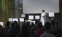 Foto vom Theaterst&uuml;ck "Diving into Math with Emmy Noether". Eine Frau in einem hellen Kleid steht auf der B&uuml;hne umringt von Schildern. Im Vordergrund sieht man das Publikum.