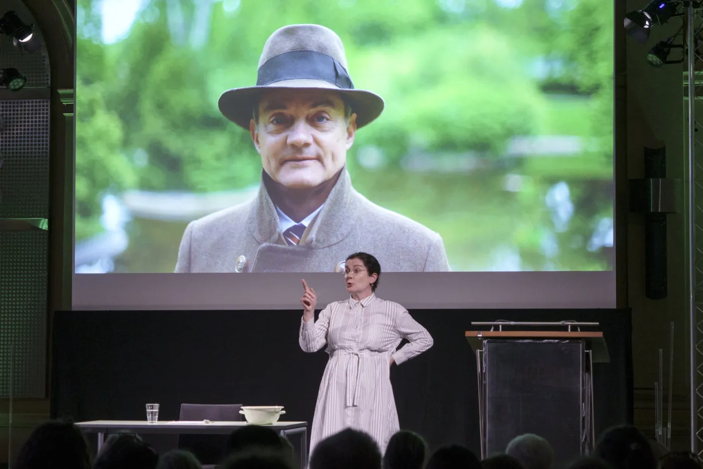 Bild vom Theaterst&uuml;ck "Diving into Math with Emmy Noether". Eine Frau in einem Kleid und runder Brille steht vor einer Leinwand. Auf der Leinwand ist ein Portrait von einem Mann mit Hut zu sehen.