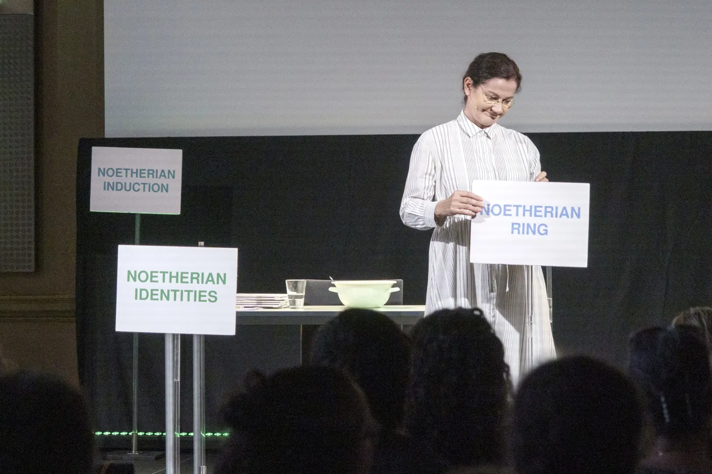 Bild vom Theaterst&uuml;ck "Diving into Math with Emmy Noether". Eine Frau in einem Kleid und runder Brille stellt Schilder auf einer B&uuml;hne auf. Darauf zu lesen: "Noetherian Ring", "Noetherian Identities", "Noetherian Induction".