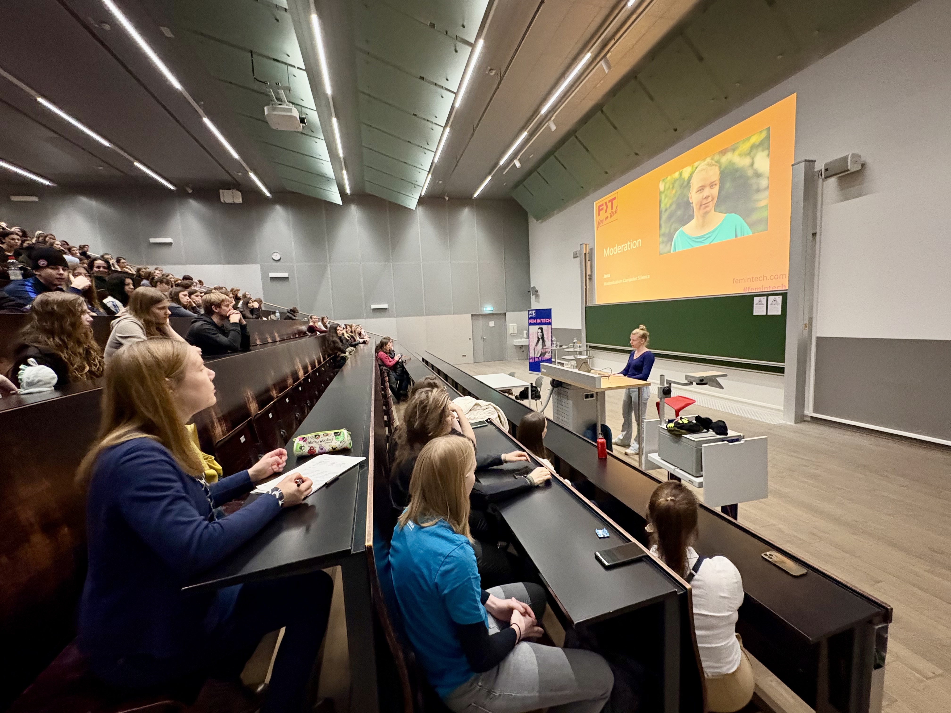Kick-Off des Fem In Tech Infotags. Moderatorin Jana steht auf der Bühne. Man sieht das Publikum in den Tribünen.