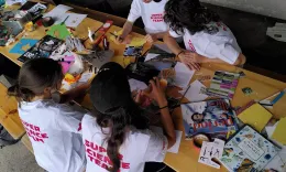 Vier Teilnehmer*innen sitzen an einem Biertisch, der mit Flyern, Magazinen und Stickern bedeckt ist. Alle tragen wei&szlig;e T-Shirts, auf denen "Super Science Team" steht.