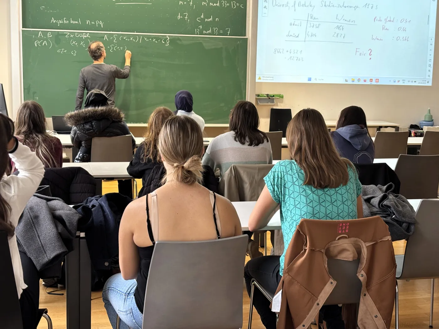 Mathematikvortrag zum Thema Wahrscheinlichkeiten: Ein Professor schreibt Formeln auf eine Tafel. Im Vordergrund sieht man Sch&uuml;lerinnen von hinten.