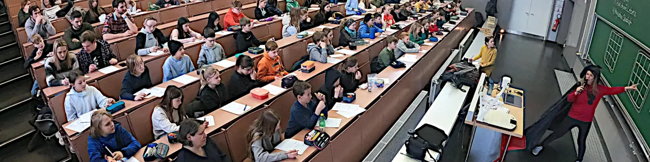 Ein Vorlesungssaal mit vielen Sch&uuml;ler*innen. Bei der Tafel steht eine Professorin, die gerade ein mathematisches Beispiel erkl&auml;rt.