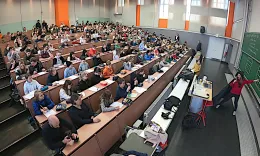 Ein Vorlesungssaal mit vielen Sch&uuml;ler*innen. Bei der Tafel steht eine Professorin, die gerade ein mathematisches Beispiel erkl&auml;rt.