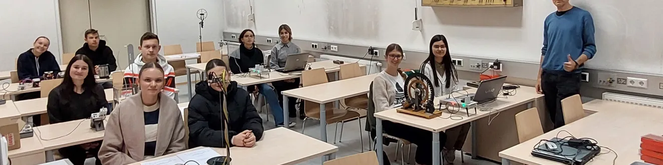 Sch&uuml;ler*innen sitzen an ihren Tischen mit Experimenten und l&auml;cheln in die Kamera.