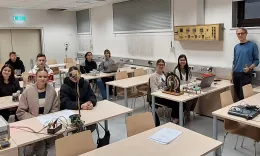 Sch&uuml;ler*innen sitzen an ihren Tischen mit Experimenten und l&auml;cheln in die Kamera.
