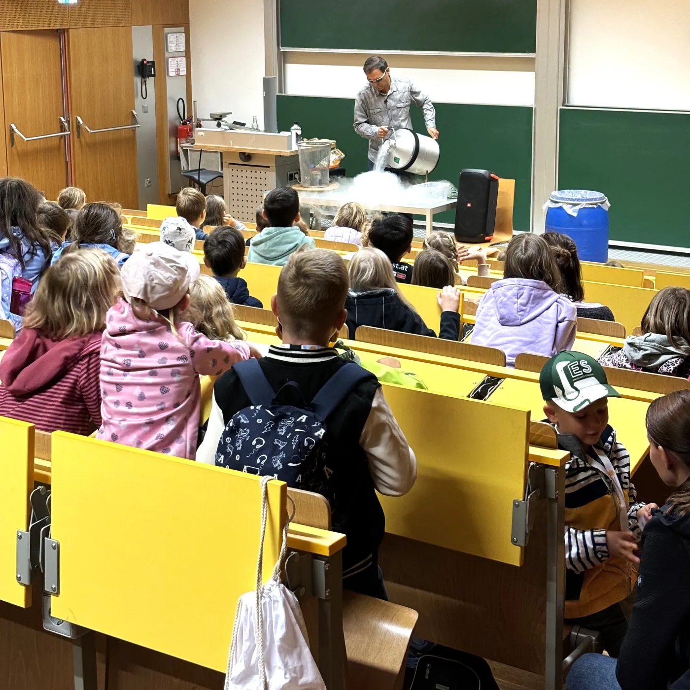 Schauvorlesung: Bernhard Weingartner steht auf auf der B&uuml;hne und sch&uuml;ttet gerade fl&uuml;ssigen Sauerstoff in ein Gef&auml;&szlig;. Der Dampf wabert &uuml;ber den Tisch. Im Publikum sitzen Kinder und Jugendliche und schauen gebannt zu.