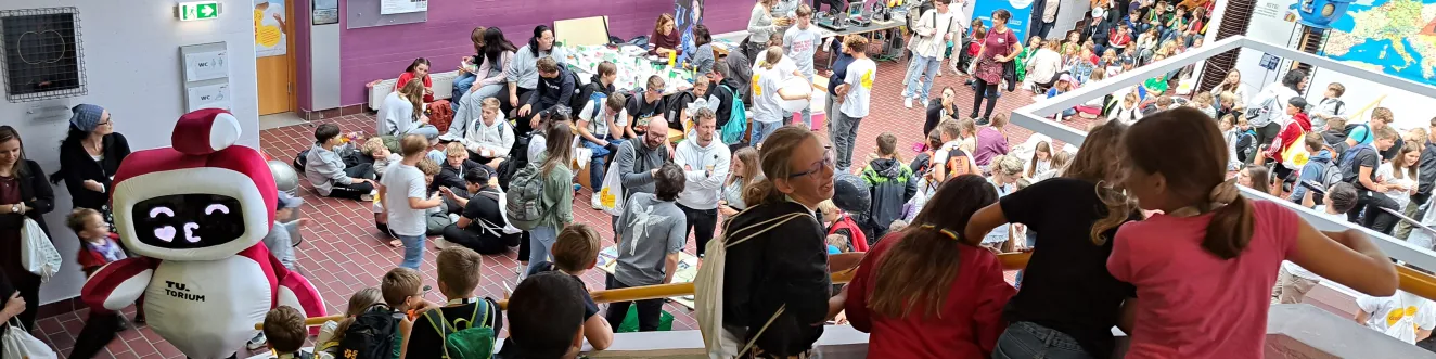 Ein dicht gef&uuml;llter Saal ist von einer erh&ouml;hten Position zu sehen. Viele Kinder und Jugendliche tummeln sich auf der Stiege und an Tischen, auf denen Experimentierstationen aufgebaut sind. In der linken Ecke sieht man ein Maskottchen - das TU.Torium - mit einem Bildschirm als Gesicht, der gerade einen Kussmund zeigt.