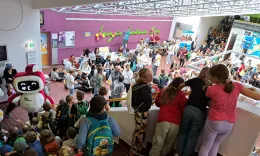 Ein dicht gef&uuml;llter Saal ist von einer erh&ouml;hten Position zu sehen. Viele Kinder und Jugendliche tummeln sich auf der Stiege und an Tischen, auf denen Experimentierstationen aufgebaut sind. In der linken Ecke sieht man ein Maskottchen - das TU.Torium - mit einem Bildschirm als Gesicht, der gerade einen Kussmund zeigt.