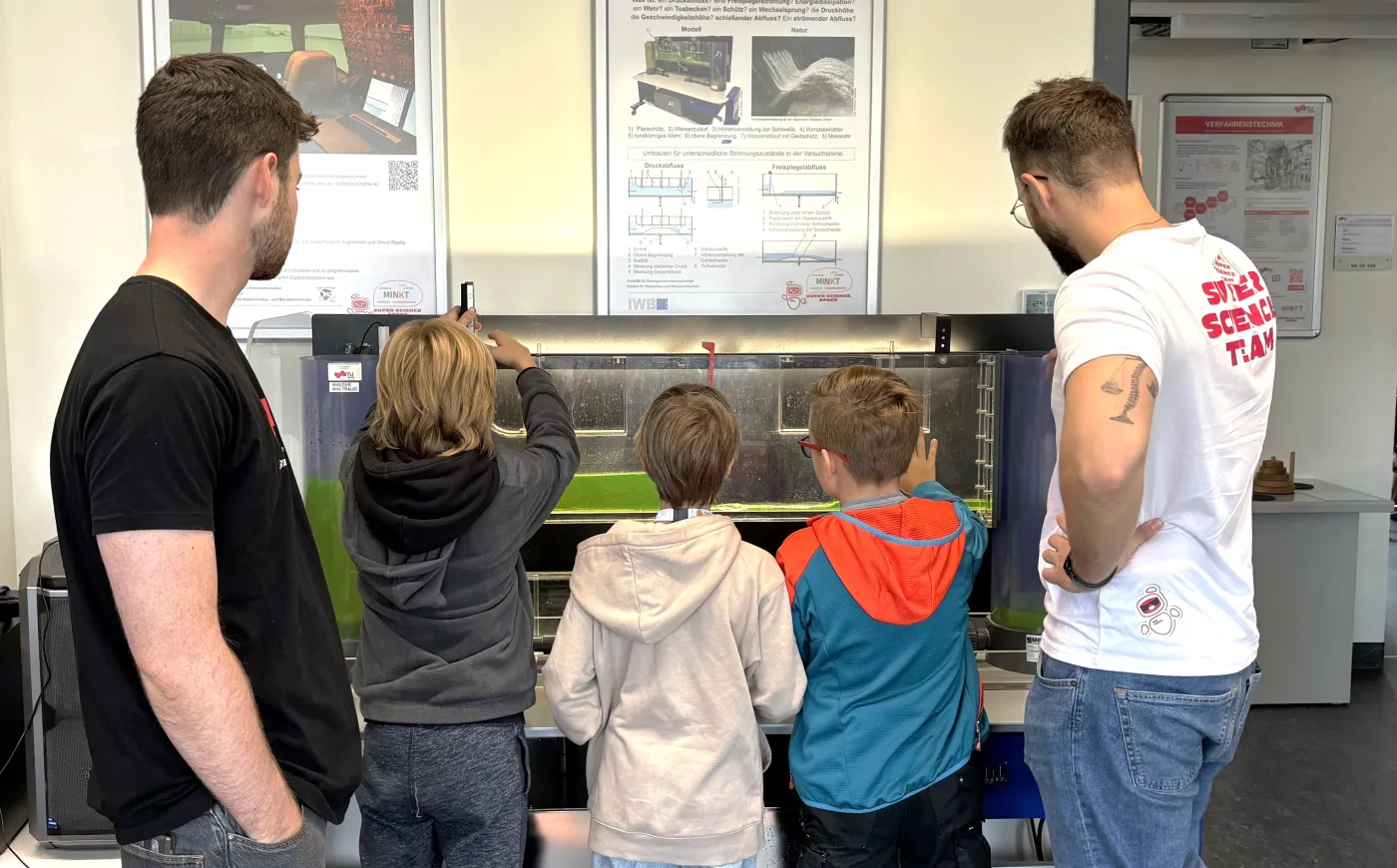 Drei Kinder und zwei Betreuer stehen vor einem Staudamm-Simulator, der mit gr&uuml;nem Wasser gef&uuml;llt ist.