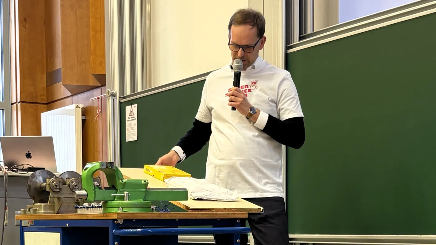 Demonstration von der Rutschf&auml;higkeit unterschiedlicher Verpackungsmaterialien in der Logistik. Zwei Pakete liegen auf einem Brett. Der Vortragende hebt das Brett auf einer Seite leicht an. Die Pakete fangen an zu rutschen.