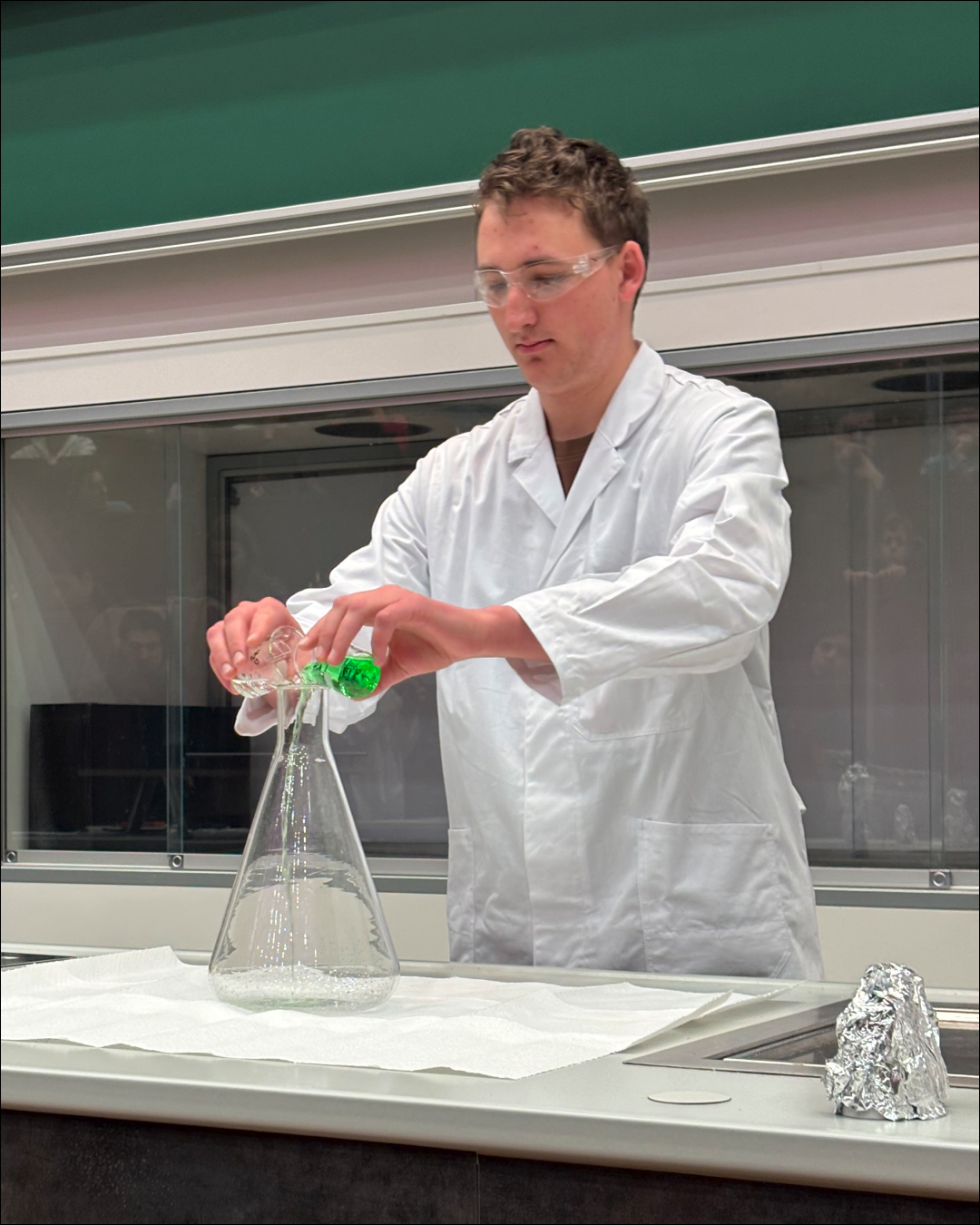 Ein Chemie-Laborant im weißen Mantel schüttet zwei unterschiedlich farbige Flüssigkeiten in einem Erlenmeyerkolben zusammen