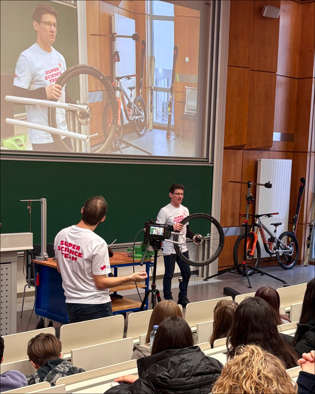 Physik-Schauvorlesung: Blick auf die Bühne. Der Lehrende erklärt gerade und hält einen Fahrradreifen in der Hand. Ein Assistent filmt.