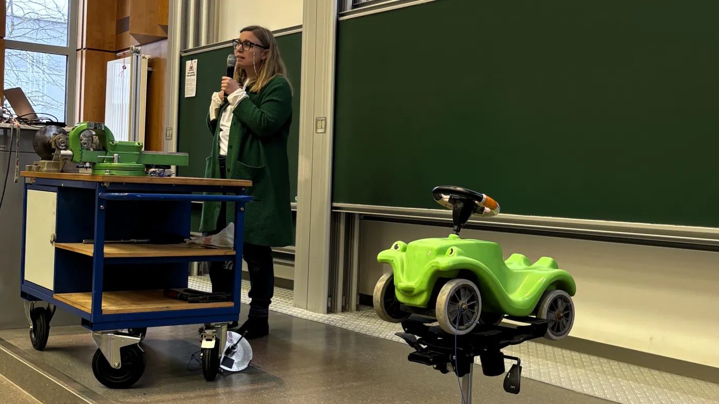 Eine Professorin tr&auml;gt vor. Im Vordergrund steht ein knallgr&uuml;nes Bobby-Car auf einem Schreibtischsessel.