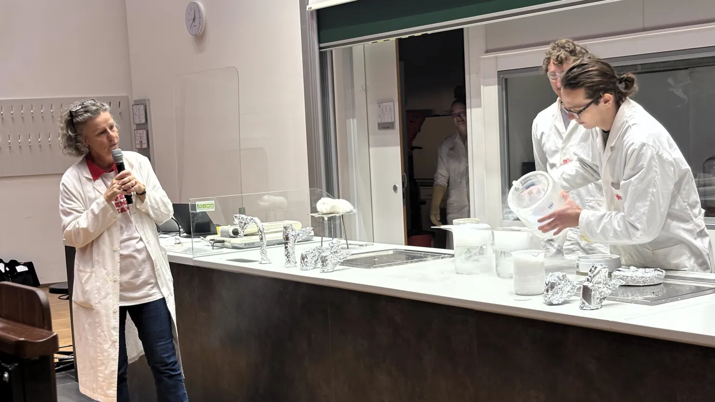 Die Professorin zeigt auf das Experiment w&auml;hrend sie vortr&auml;gt. Ihre beiden Assistent*innen gie&szlig;en eine dampfende Fl&uuml;ssigkeit in ein Beh&auml;ltnis.