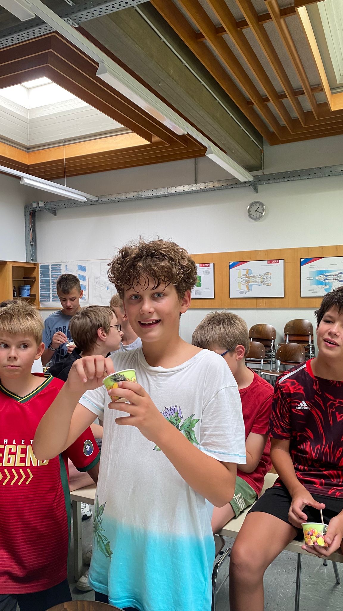 Ein Bub mit gelockten Haaren l&auml;chelt in die Kamera. Er h&auml;lt einen Eiscreme-Becher in der Hand. Im Hintergrund sieht man viele andere Kinder, die ebenfalls ein Eis genie&szlig;en.