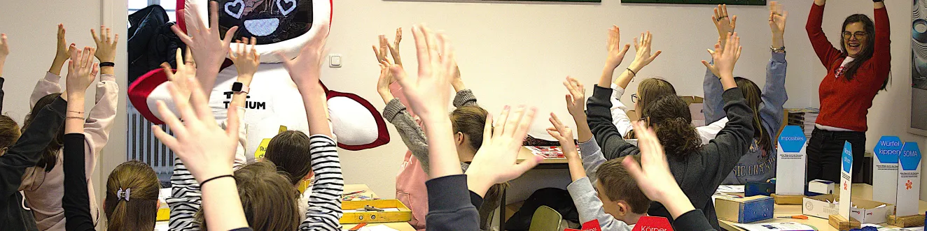 Das TU.Torium ist da! Das Roboter-Maskottchen mit Herzaugen winkt der Klasse zu. Die Sch&uuml;ler*innen strecken erfreut die Arme in die H&ouml;he.