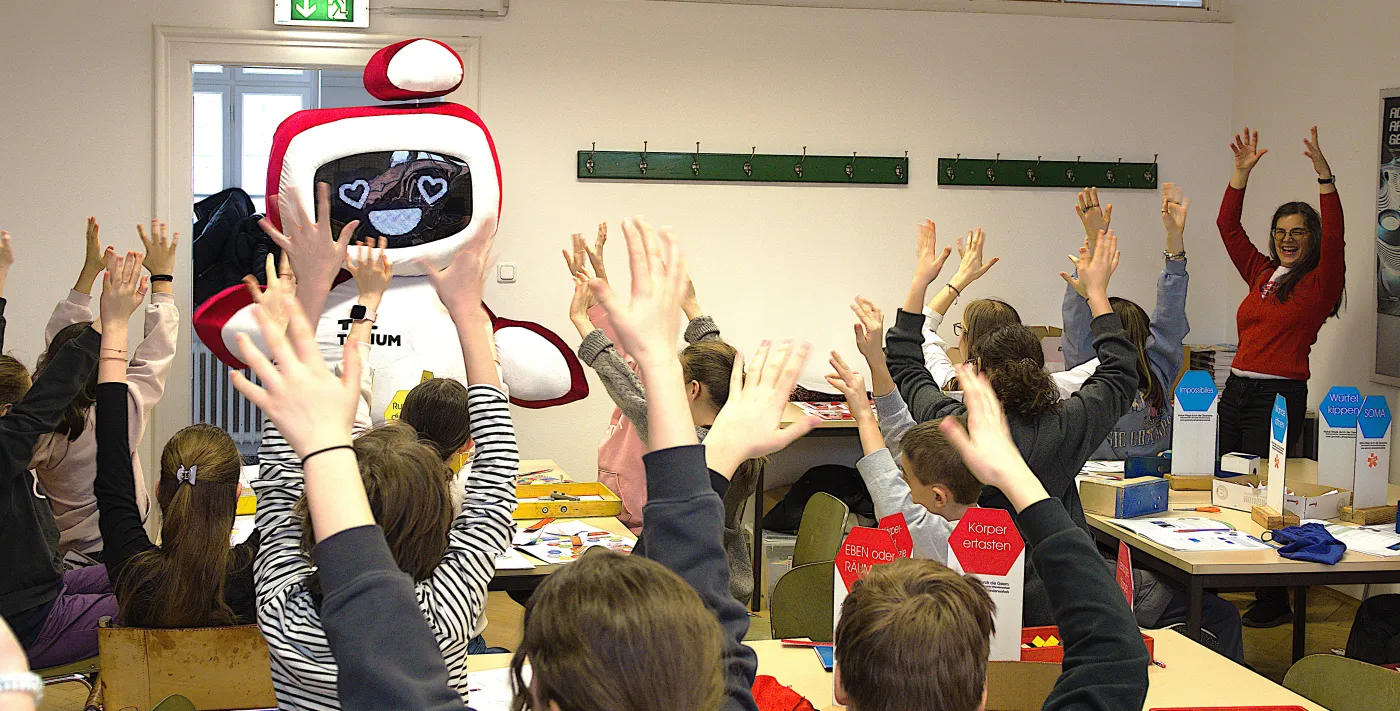 Das TU.Torium ist da! Das Roboter-Maskottchen mit Herzaugen winkt der Klasse zu. Die Sch&uuml;ler*innen strecken erfreut die Arme in die H&ouml;he.