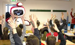 Das TU.Torium ist da! Das Roboter-Maskottchen mit Herzaugen winkt der Klasse zu. Die Sch&uuml;ler*innen strecken erfreut die Arme in die H&ouml;he.