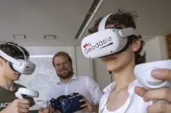 2 Jugendliche tragen Virtual Reality Brillen und jeweils zwei Controller. Auf den Brillen steht das Wort Geodäsie unter dem TU Graz Logo.